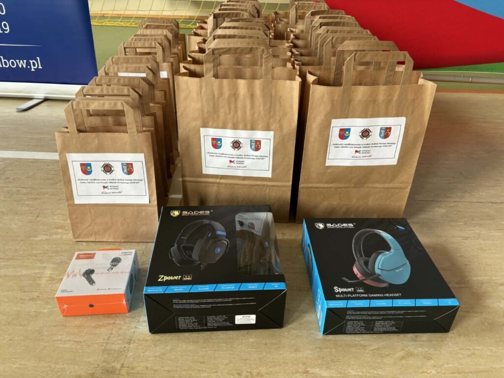 Zdjęcie przedstawia rząd brązowych papierowych toreb z nadrukowanymi emblemami. Przed nimi są pudełka ze słuchawkami w czarnym i niebieskim kolorze. Tło ma sportowe linie i czerwoną siatkę.