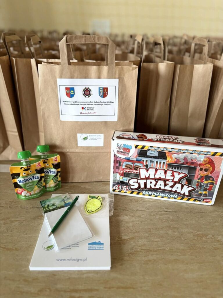 Obraz przedstawia zestaw upominkowy złożony z brązowych toreb, gry planszowej Mały Strażak, soczków Bobovita, notesu i długopisu. Tło jest rozmyte, co sugeruje wnętrze pomieszczenia z dalszymi toreb