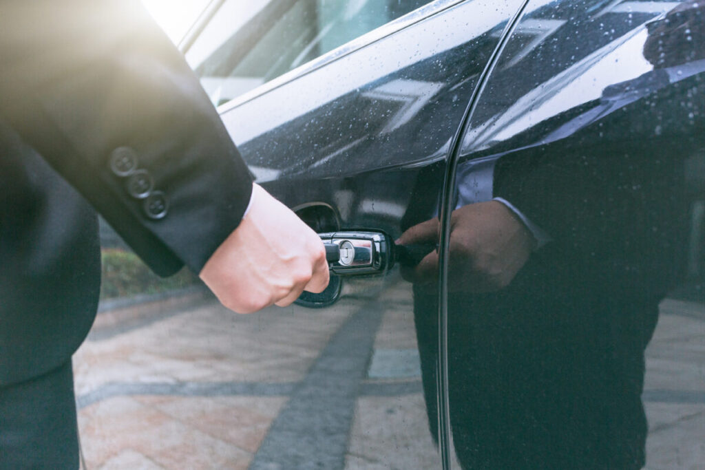 Osoba w garniturze otwiera ciemne drzwi samochodu. Słoneczne światło odbija się na lakierze. W tle widać brukowany chodnik.