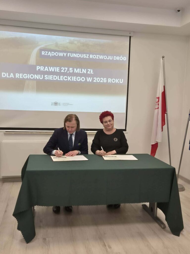 Dwóch ludzi podpisuje dokumenty przy zielonym stole. W tle ekran z napisem o funduszu drogowym na 2026 rok. Obok stoi flaga Polski.