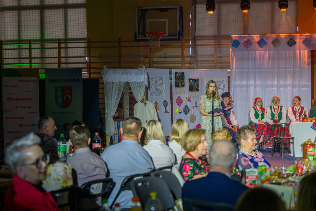 Na scenie kobieta w zielonej sukience przemawia do mikrofonu. Obok członkowie zespołu w strojach ludowych siedzą przy stole. Tło dekorowane jest materiałami w etniczne wzory i delikatnym oświetleniem.