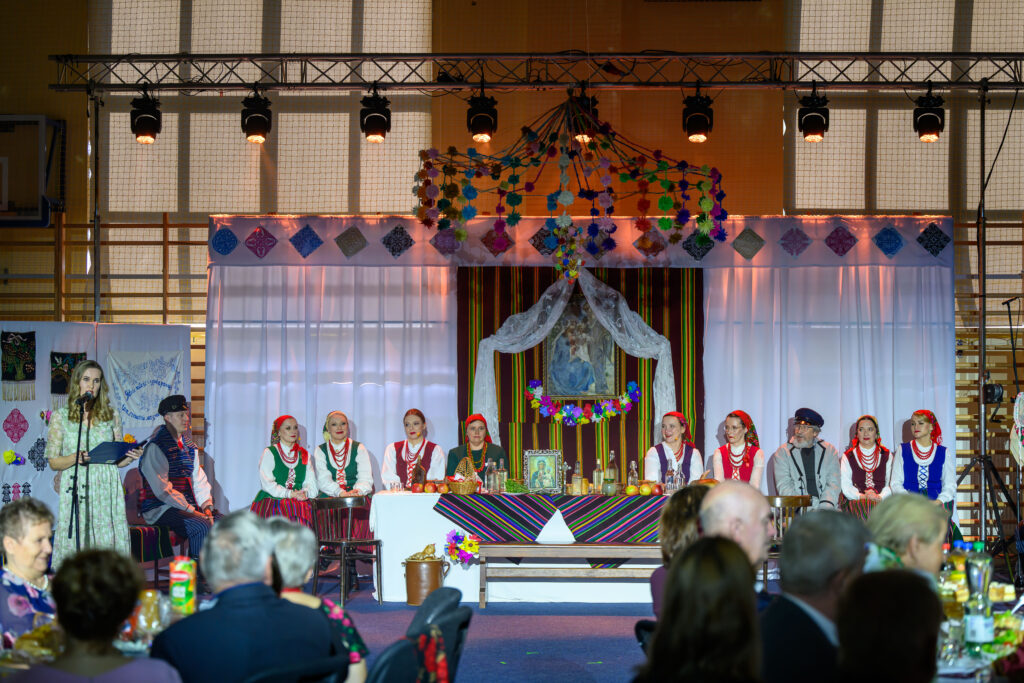 Grupa ludzi w ludowych strojach siedzi przy udekorowanym stole na scenie. Ściana w tle jest ozdobiona kolorowymi tkaninami i kwiatami. Oświetlenie sceniczne podkreśla występ.