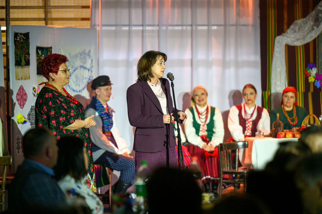 Grupa w tradycyjnych strojach ludowych na scenie. W centrum kobieta przemawia do mikrofonu, z tyłu stół z owocami. Tło zdobią hafty i kolorowe paski.