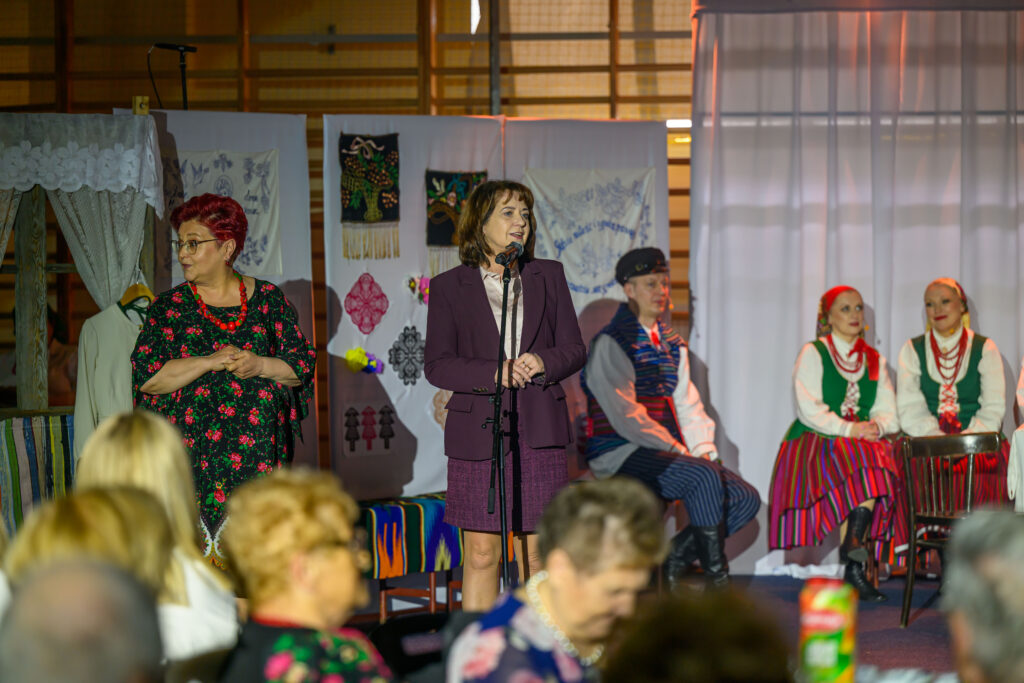 Na scenie stoi kobieta przy mikrofonie, w tle ludzie w strojach ludowych. Dekoracje w jasnych kolorach. Publiczność siedzi z przodu.