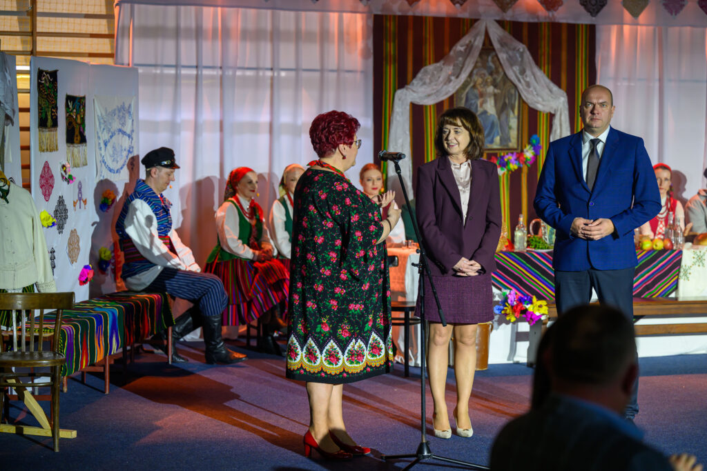 Wnętrze z kolorowymi, ludowymi strojami. Dwie osoby przemawiają do mikrofonu. W tle siedzą postacie w tradycyjnych ubraniach.