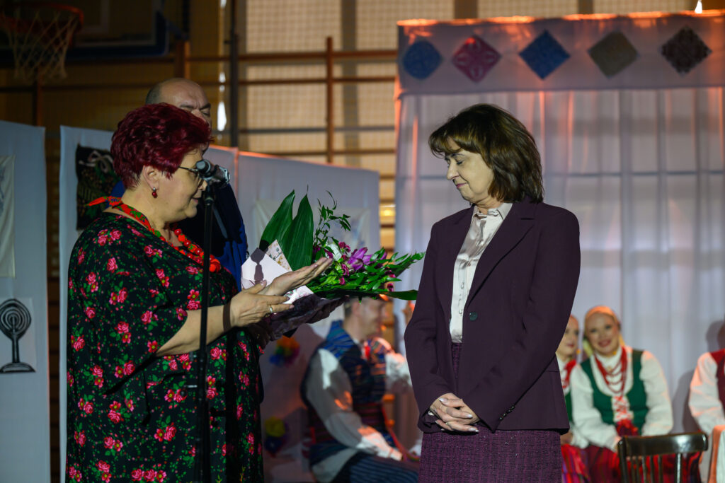 Dwie kobiety na scenie; jedna wręcza bukiet kwiatów. Tło zdobią jasne zasłony i uśmiechnięci ludzie w strojach ludowych. Kolory: zielony, fioletowy, czerwony.