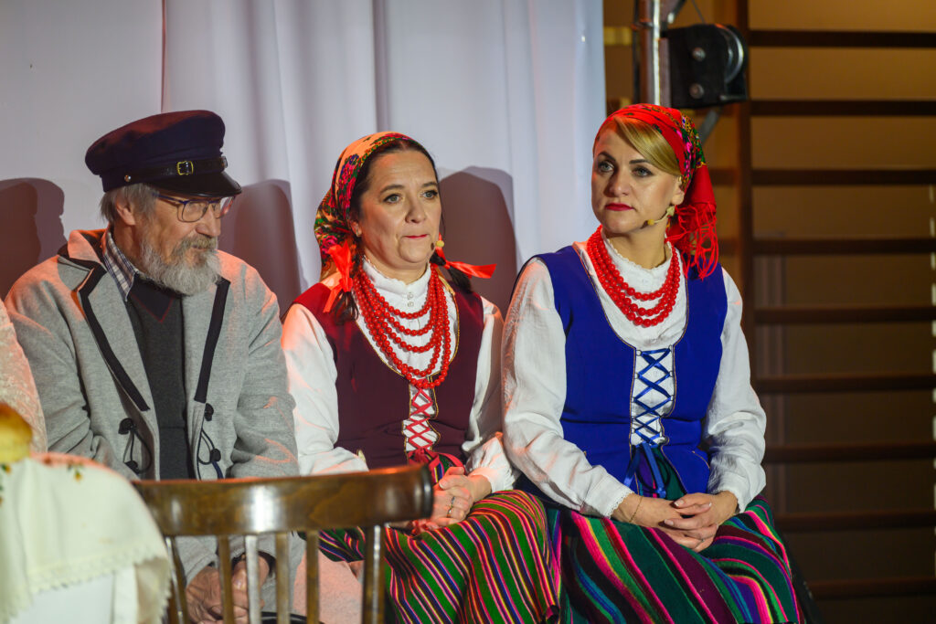 Trzy osoby w strojach ludowych siedzą na krzesłach. Dwie kobiety mają kolorowe spódnice i korale, mężczyzna nosi czapkę i okulary. Tło jest jasne, zasłonięte białą kotarą.