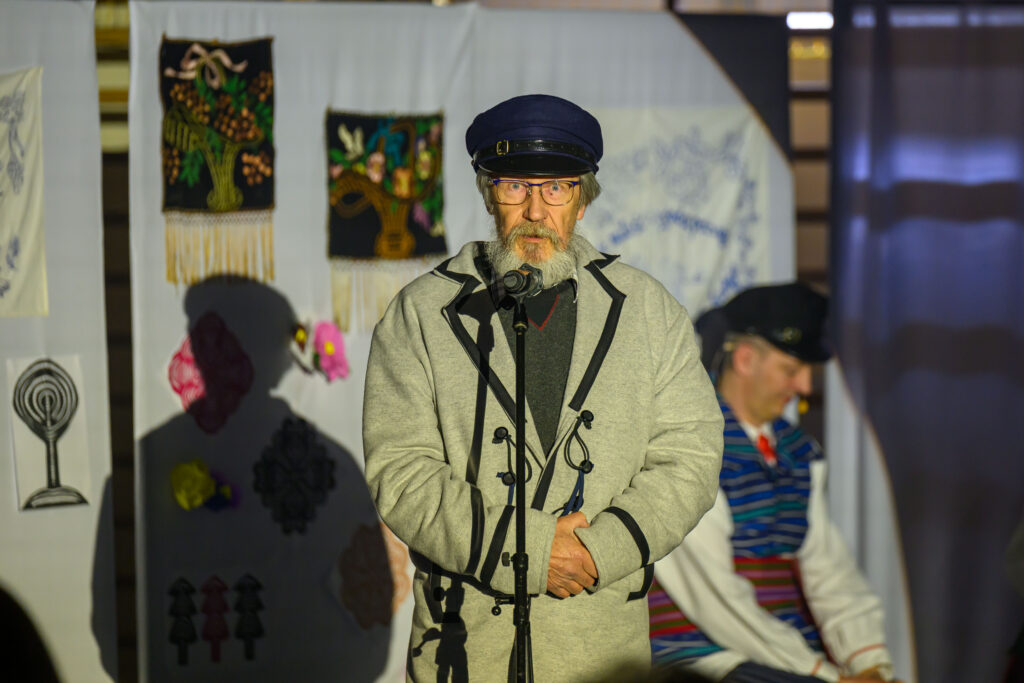 Starszy mężczyzna w szarym płaszczu i granatowej czapce stoi przy mikrofonie. W tle tkaniny z kolorowymi wzorami. Scena oświetlona jasnym światłem.