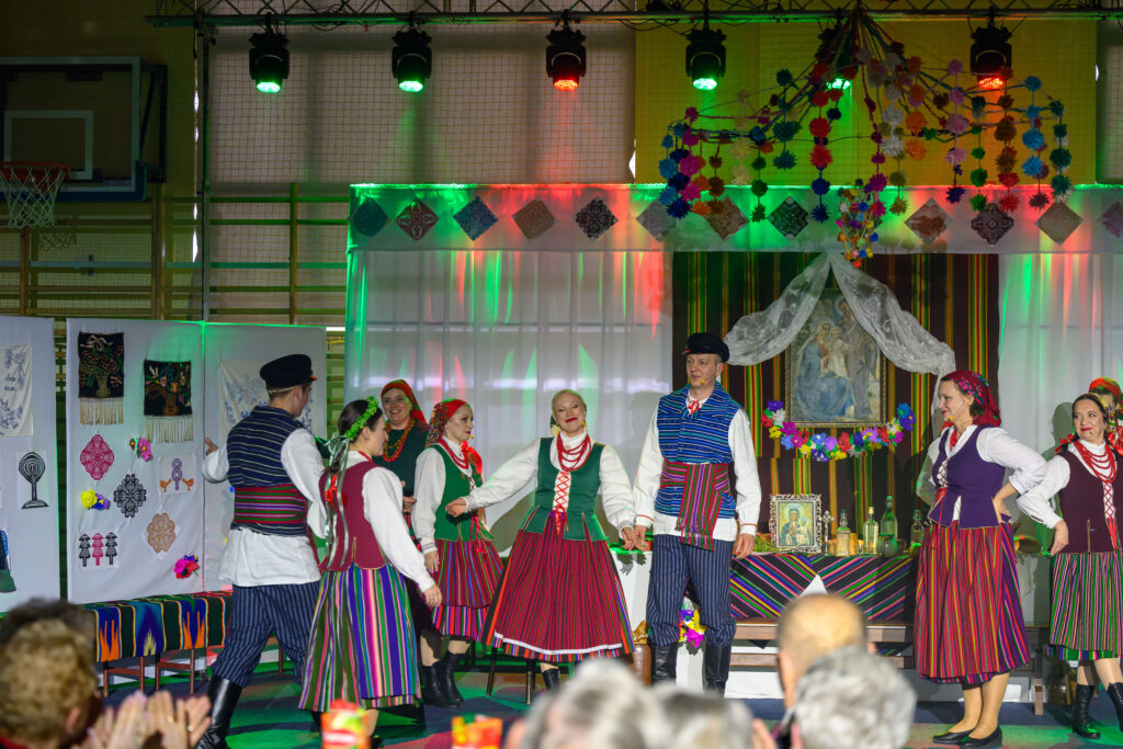 Grupa ludzi w kolorowych, ludowych strojach tańczy na scenie. Tło zdobią barwne dekoracje i oświetlenie. Scena jest jasna i radosna.