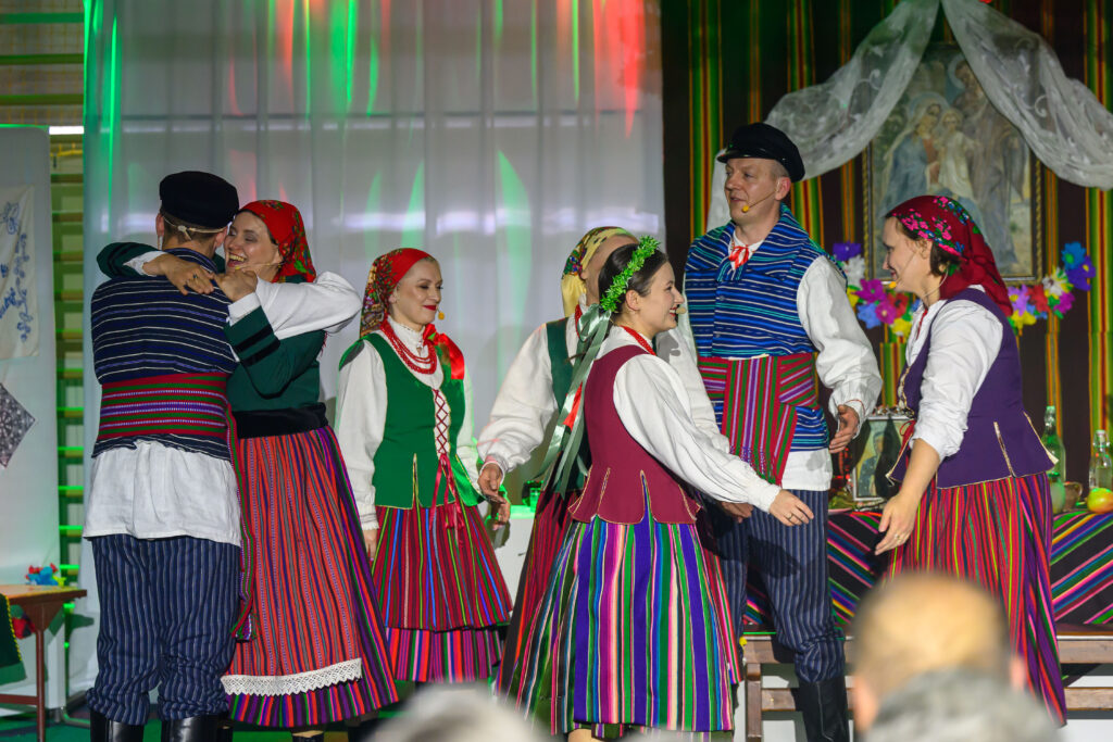 Grupa osób w strojach ludowych tańczy; dominują kolory zielony, czerwony i niebieski. Tło zdobią zasłony i obraz, a światła sceniczne tworzą kolorową aurę. W tle widoczne są kolorowe kwiaty i tradycyjne dekoracje.