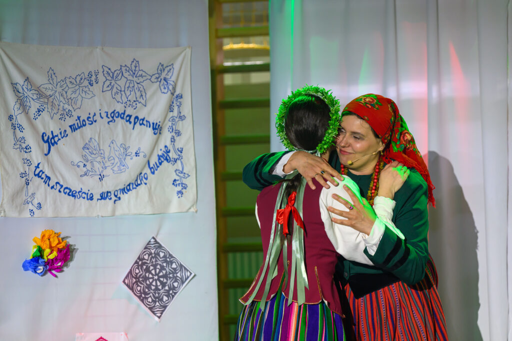 Dwie osoby w tradycyjnych, kolorowych strojach ludowych obejmują się na tle haftowanej płachty z napisem.