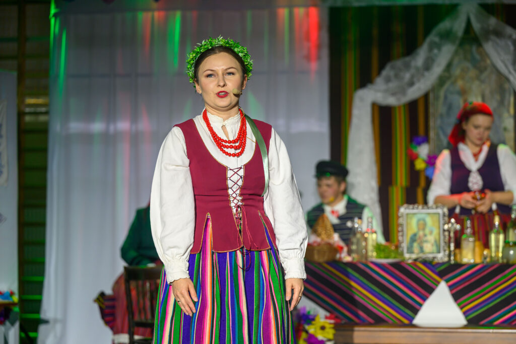 Kobieta w stroju ludowym z wiankiem i koralami na scenie. Tło z tkaninami i stołem pełnym kolorowych przedmiotów. Obok inni w tradycyjnych strojach.