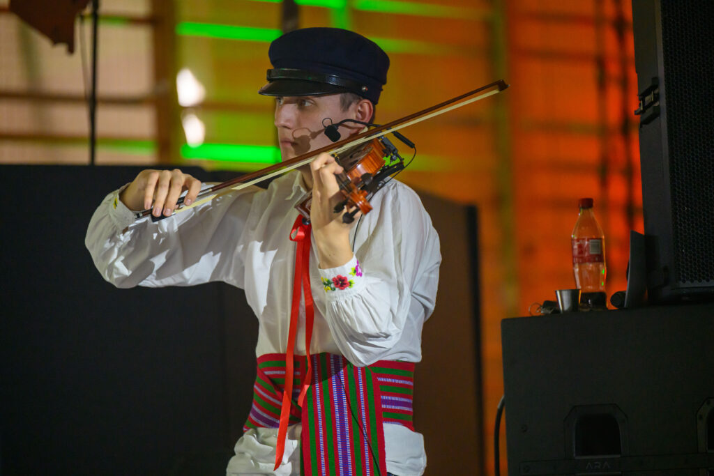 Mężczyzna w białej koszuli gra na skrzypcach, ma kolorową wstążkę i czarną czapkę. Tło jest oświetlone na zielono i pomarańczowo. Obok stoi butelka napoju.