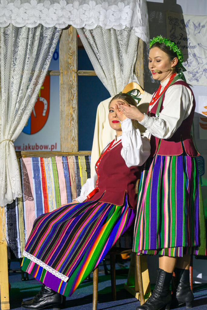 Dwie kobiety w tradycyjnych strojach ludowych w pasiaste kolory. Jedna siedzi, druga stoi obok, w tle koronkowe zasłony. Klarowne oświetlenie.