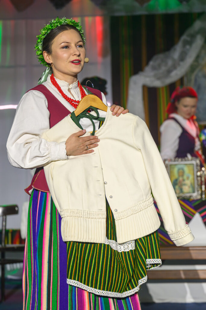 Osoba w stroju ludowym trzyma biały żakiet. Tło kolorowe z pionowymi pasami. Zielony wianek na głowie dodaje szczegółu.