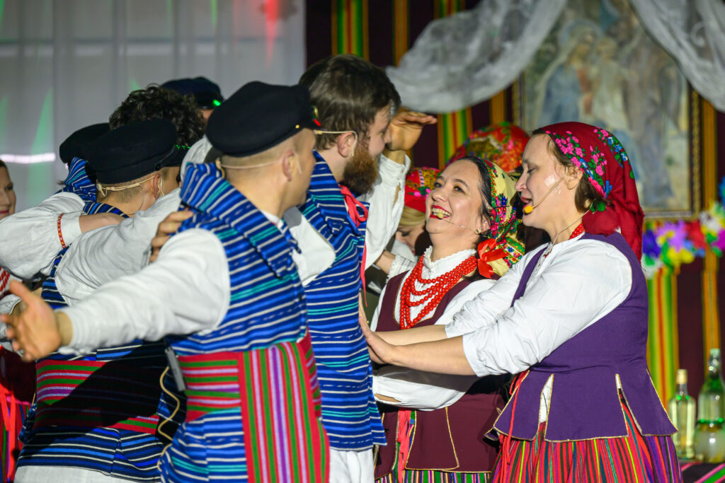 Grupa tancerzy w kolorowych strojach ludowych tańczy radośnie. Dominują odcienie niebieskiego, czerwonego i fioletu. W tle zawieszone są kolorowe dekoracje.