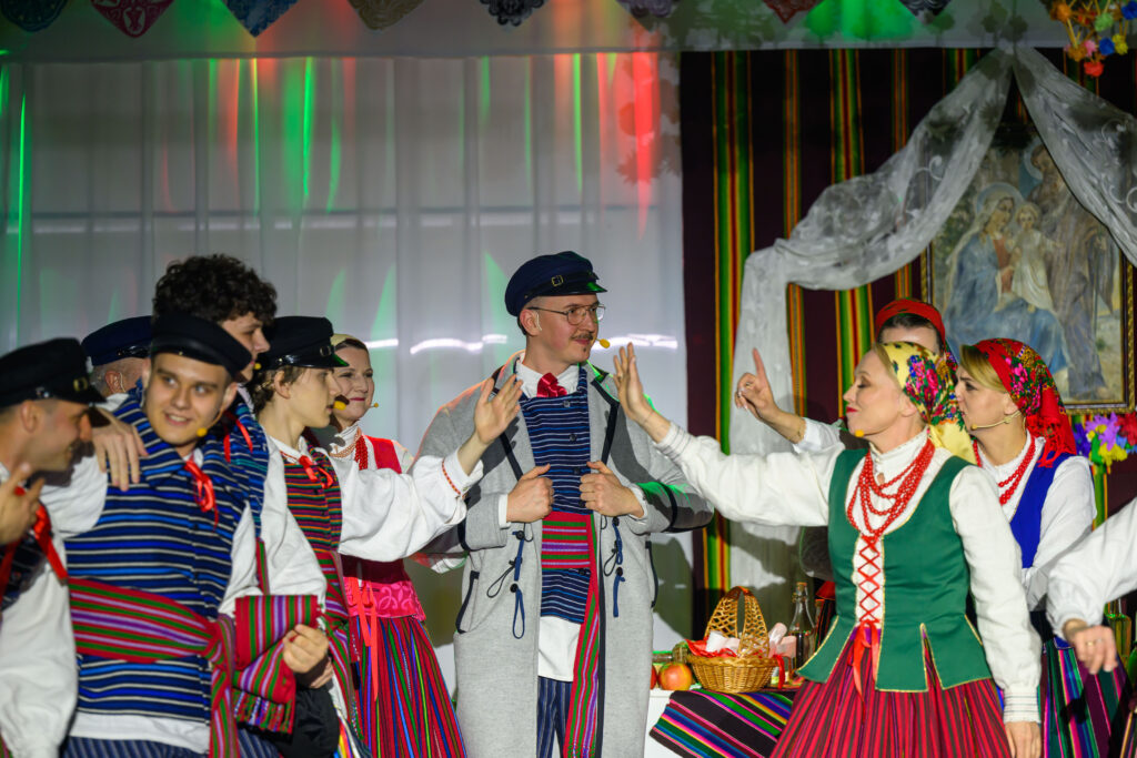 Grupa ludzi ubranych w kolorowe, tradycyjne stroje ludowe tańczy na scenie. Tło jest jasne z zielonymi i czerwonymi światłami. W tle widać kosz piknikowy i obraz.