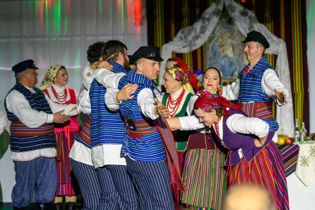 Tradycyjny taniec w kolorowych strojach ludowych. Dominują pasy w kolorach: niebieski, czerwony, zielony i fioletowy. Tło to jasna ściana z dekoracją.