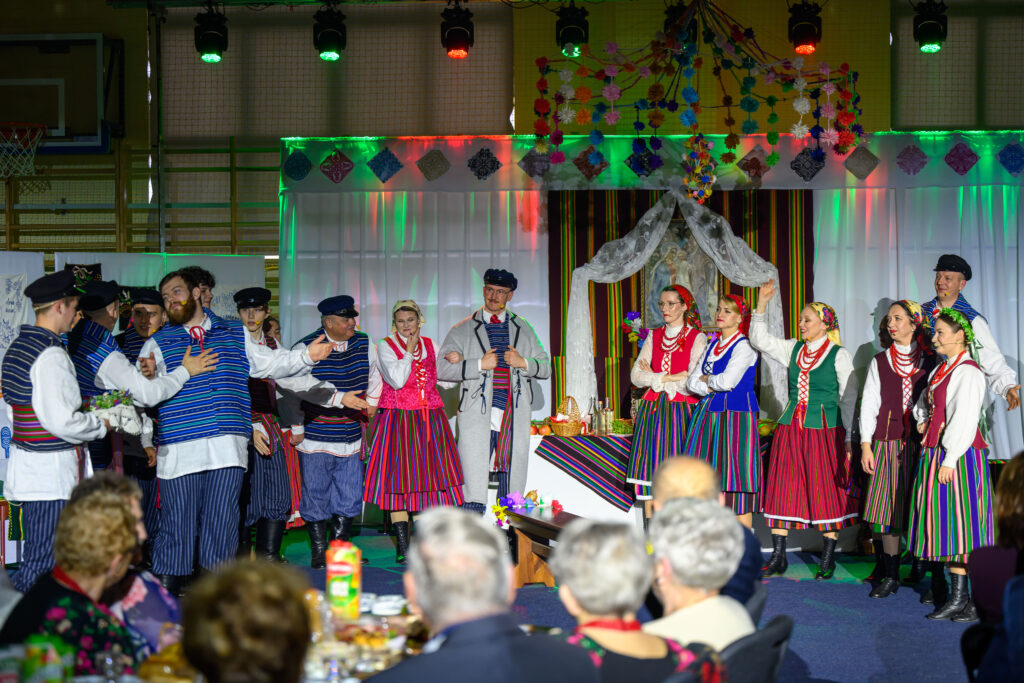 Grupa ludzi w kolorowych strojach ludowych tańczy na scenie. W tle dekoracje z kwiatami, a scena jest oświetlona na zielono i czerwono. Publiczność widoczna z przodu.