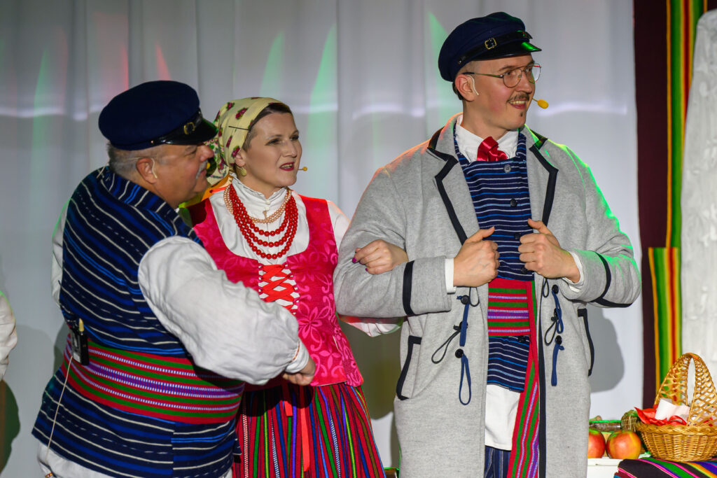 Trzy osoby w strojach ludowych na scenie. Jaskrawe kolory: różowy, niebieski, szary. Koszyk z jabłkami w tle.