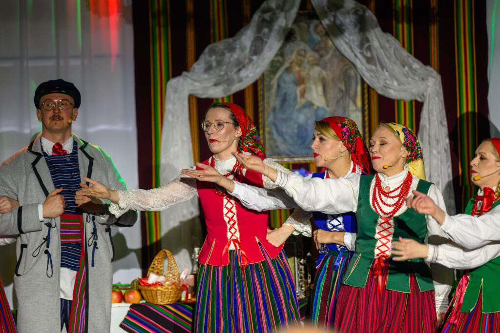 Grupa osób w kolorowych strojach ludowych tańczy na scenie. Dominuje czerwień, zieleń i różne odcienie pasków. W tle obraz i kosz z owocami na stole.