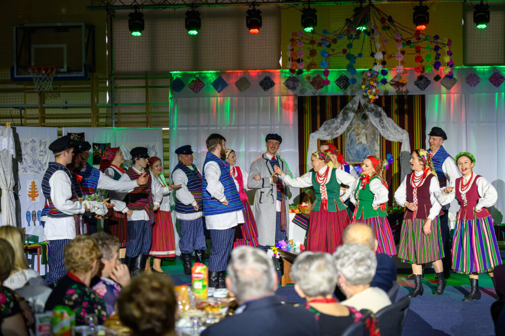 Grupa osób w tradycyjnych strojach ludowych tańczy na scenie. Tło jest udekorowane kolorowymi światłami i ozdobami. Widzowie siedzą przy stole z jedzeniem.