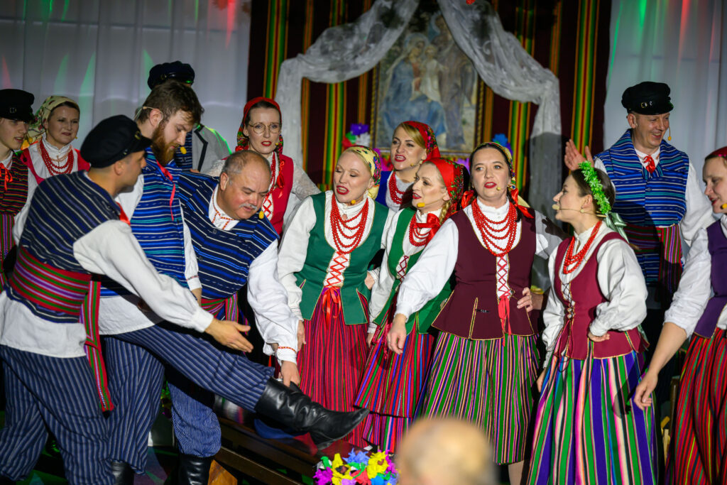 Grupa ludzi w kolorowych strojach ludowych tańczy na scenie. Tło ozdobione jest materiałami i obrazem. Dominują kolory czerwony, zielony i niebieski.