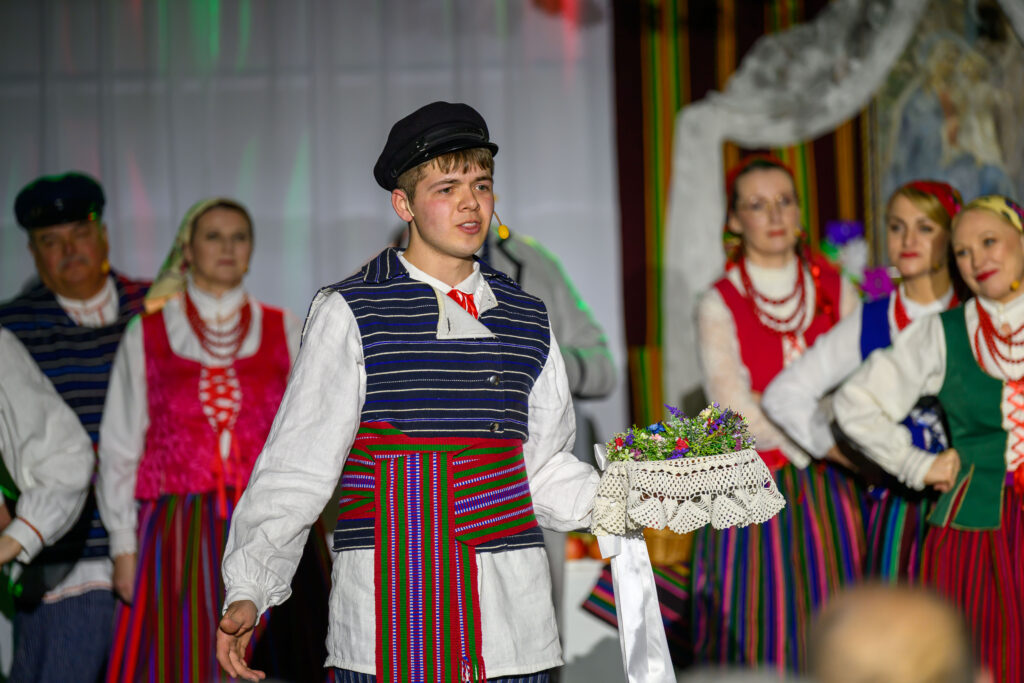 Grupa osób w ludowych strojach o żywych kolorach na scenie, trzymająca koszyk z kwiatami. Kolorowe wstęgi i lamówka. Tło sceniczne jest rozmyte.