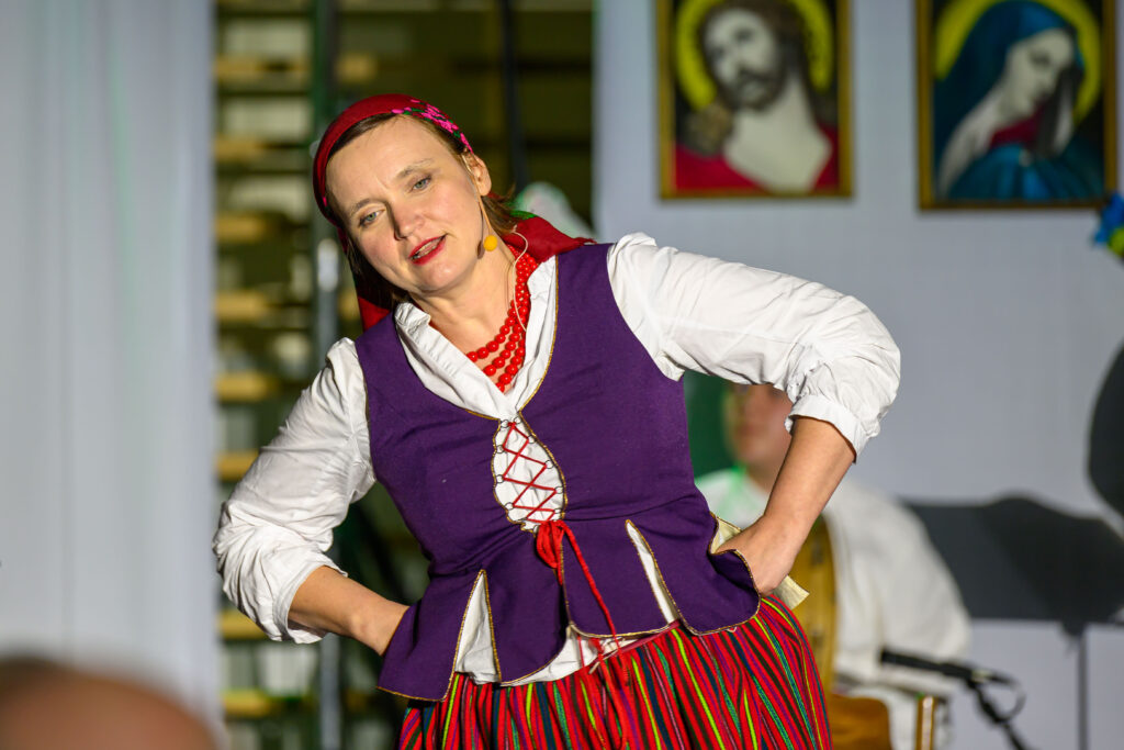 Kobieta w tradycyjnym stroju ludowym, z fioletową kamizelką i czerwoną spódnicą, stoi na scenie. W tle widać obrazy religijne. Oświetlenie skupia się na postaci, reszta jest rozmyta.