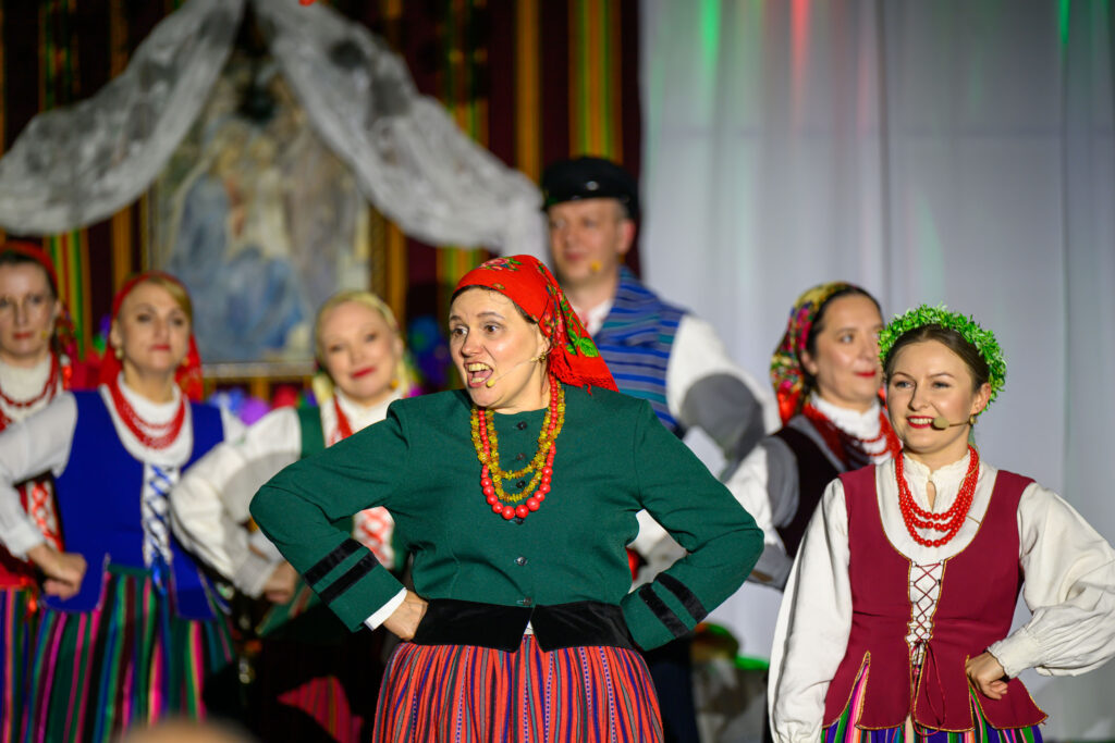 Grupa osób w kolorowych, tradycyjnych polskich strojach ludowych, stoi na scenie. Tło jest zdobione białymi zasłonami i obrazem. Dominują kolory czerwony, zielony i niebieski.