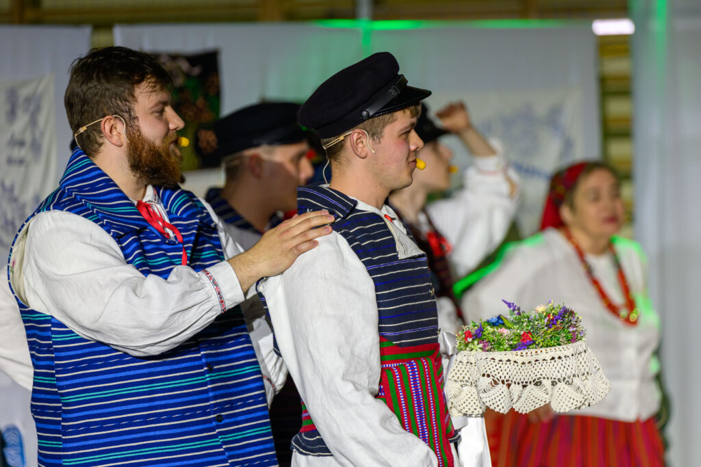 Grupa ludzi w tradycyjnych strojach ludowych tańczy na scenie. Ubrania w niebieskie i czerwone paski kontrastują z białym tłem. W rękach trzymają kosz z kwiatami.