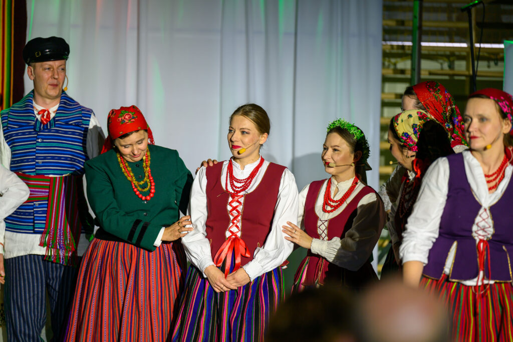 Grupa osób w strojach ludowych na tle białej zasłony. Kolorowe ubrania w paski i kwiatowe wzory. Ciepłe oświetlenie sceniczne.