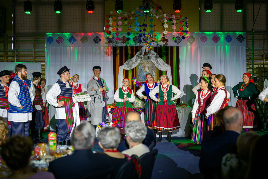 Grupa osób w tradycyjnych strojach ludowych tańczy na scenie. Kolorowe stroje i dekoracje tworzą tło. Publiczność siedzi na pierwszym planie.