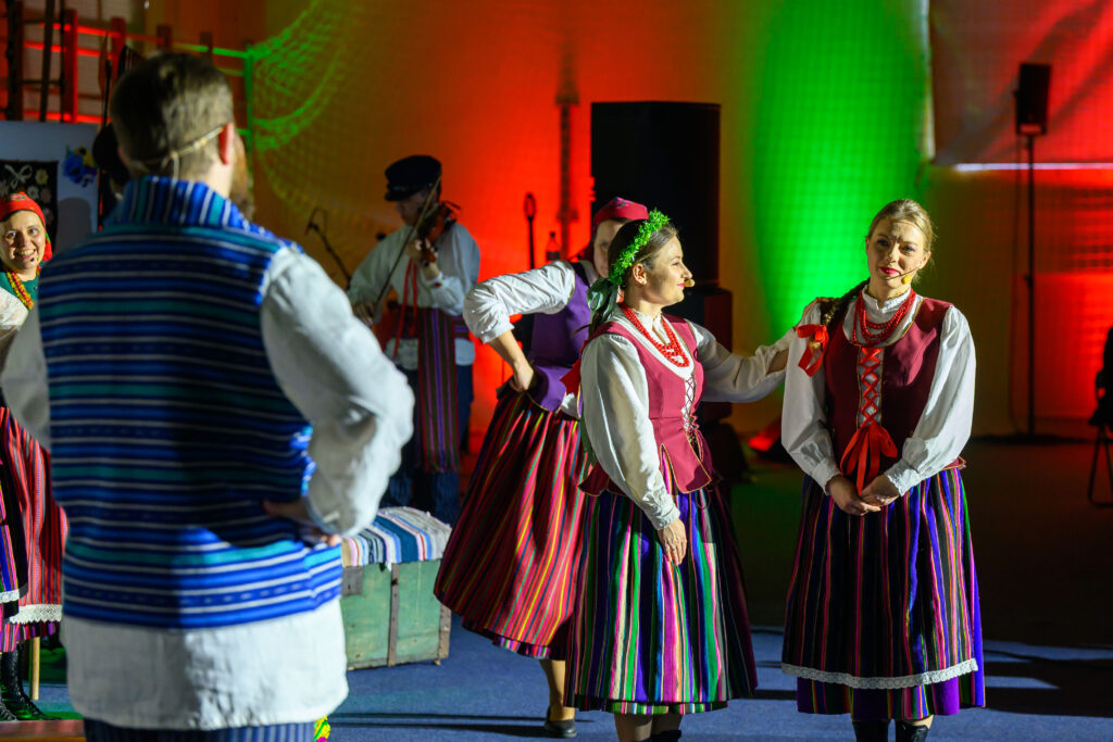 Ludzie w strojach ludowych tańczą na scenie. Kolorowe światła w tle, głównie czerwone i zielone. Stroje mają pasiaste wzory i białe koszule.