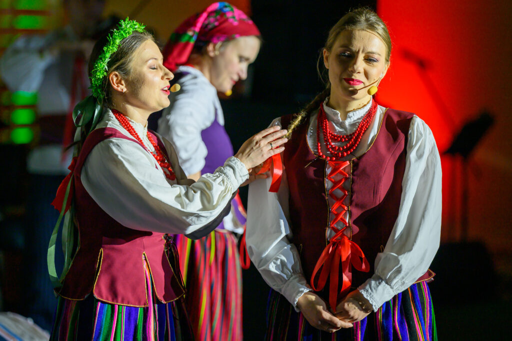 Kobiety w strojach ludowych śpiewają na scenie. Mają kolorowe spódnice i czerwone korale. Tło ciemne z ciepłym oświetleniem.