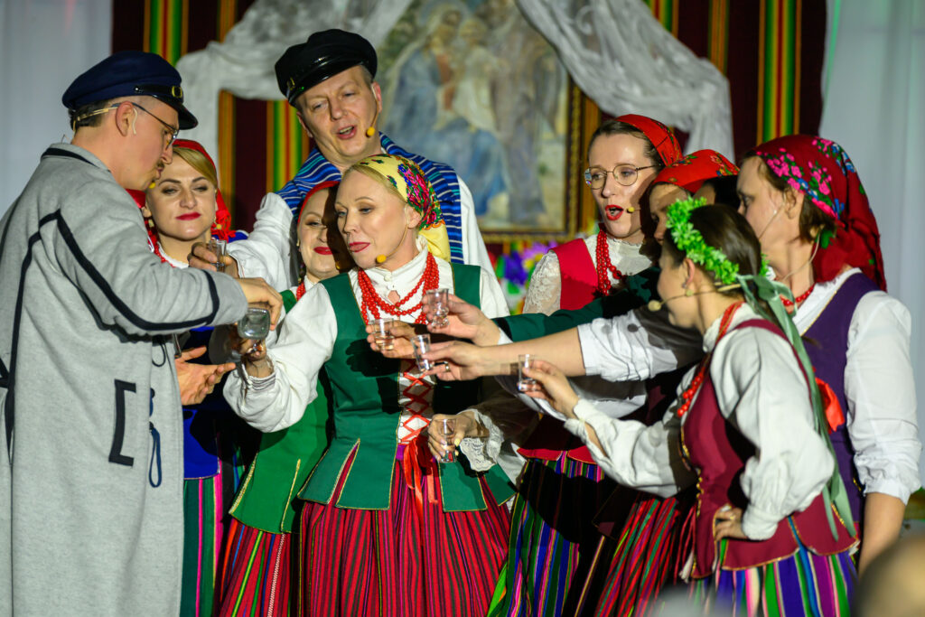 Grupa ludzi w kolorowych, tradycyjnych strojach, wesoło toastingu. Przeważają zielenie i czerwienie. W tle ozdobne tkaniny i obraz.