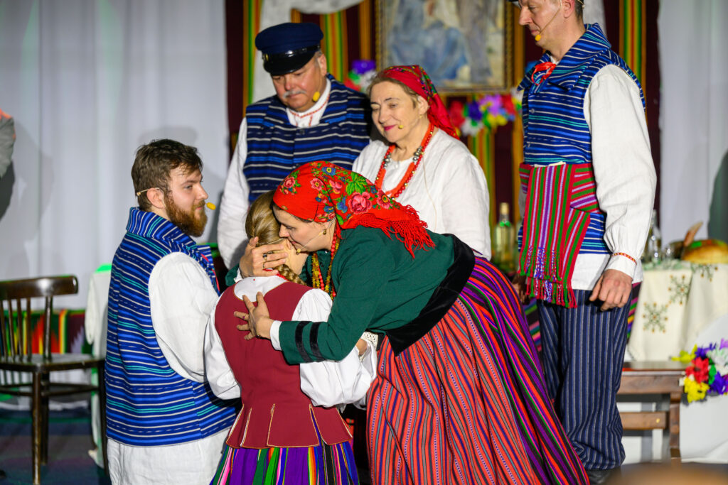 Tradycyjnie ubrani ludzie w barwnych, ludowych strojach nawiązują interakcję na scenie. Tło zdobi wzorzysty materiał. Atmosfera jest żywa i barwna.