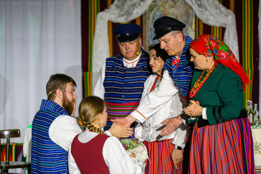 Grupa osób w kolorowych strojach ludowych wykonuje tradycyjny obrzęd. Dominuje niebieski, czerwony i zielony strój. Tło ozdobne z białymi zasłonami.