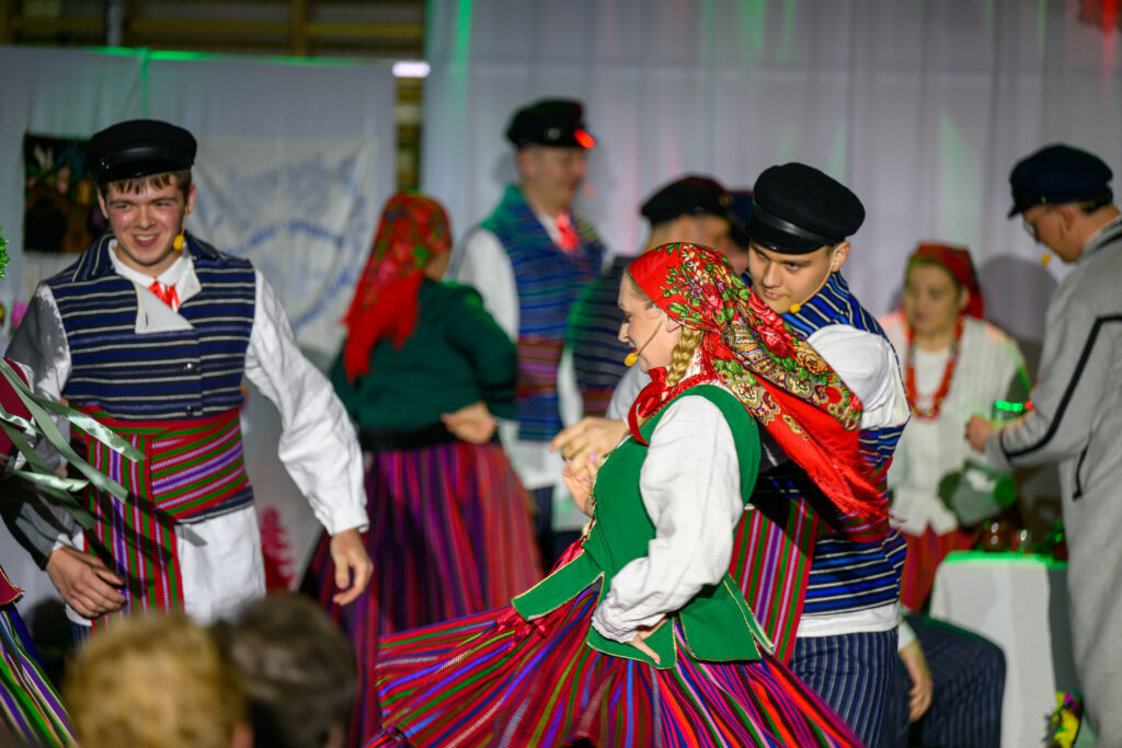 Taniec ludowy na scenie, kolorowe stroje ludowe, czerwone i zielone akcenty; w tle biała dekoracja i tańczący ludzie.