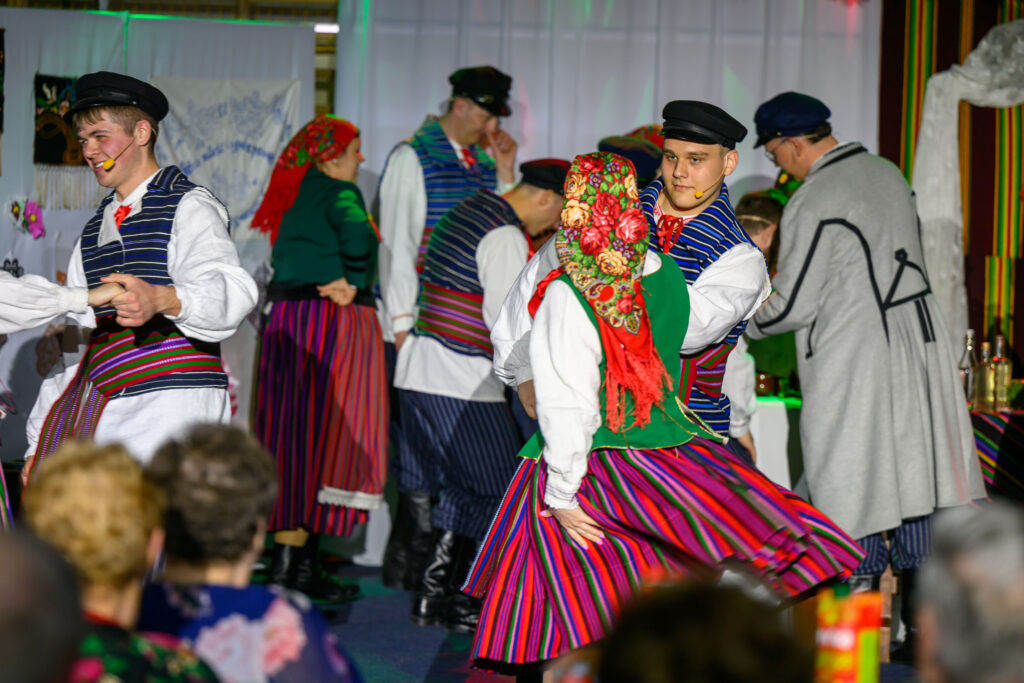 Ludzie w tradycyjnych, kolorowych strojach ludowych tańczą na scenie. Dominują kolory czerwony, zielony i niebieski. W tle biały ekran i tkaniny dekoracyjne.