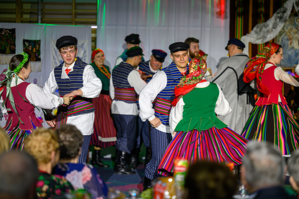 Ludzie w kolorowych, tradycyjnych strojach tańczą na scenie. Scena jest jasno oświetlona. W tle białe zasłony.