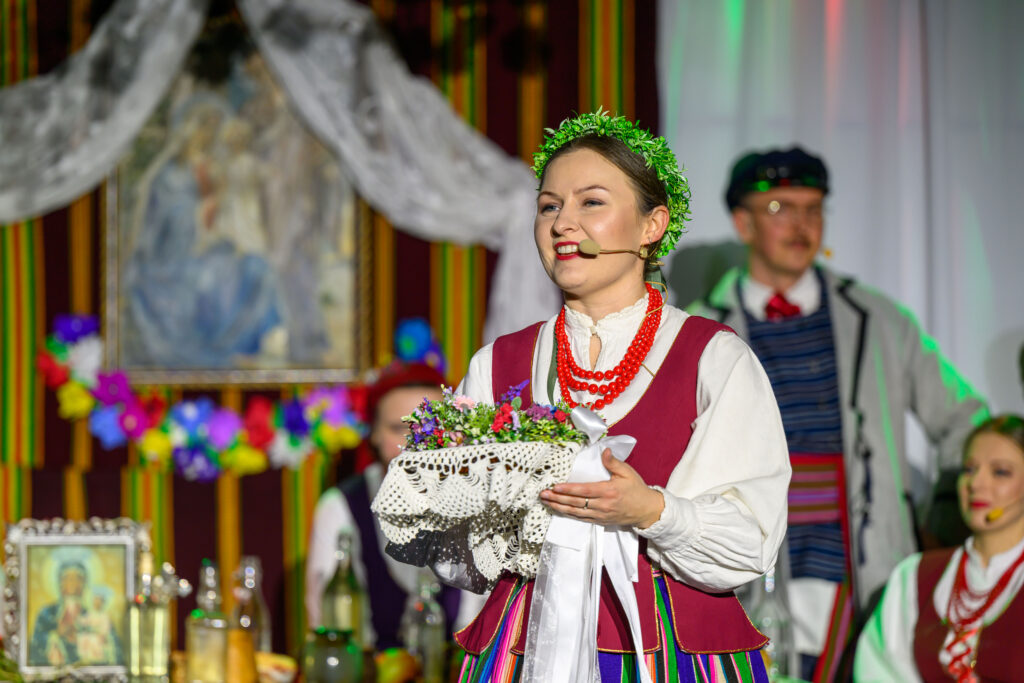 Kobieta w kolorowym, ludowym stroju trzyma kosz z kwiatami. Na tle obraz i dekoracje w jasnych barwach. Widać również inne osoby.