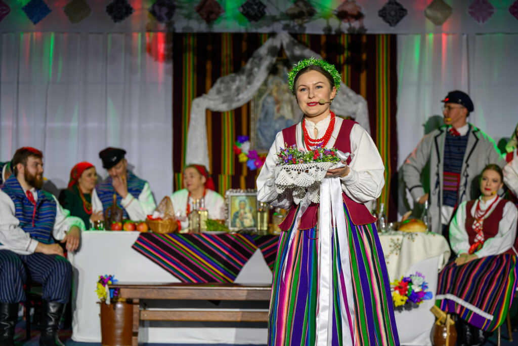 Na scenie kobieta w kolorowym, ludowym stroju trzyma koszyk z kwiatami. Za nią znajduje się stół z ludźmi w tradycyjnych ubraniach. Tło jest ozdobione barwnymi materiałami i światłami.