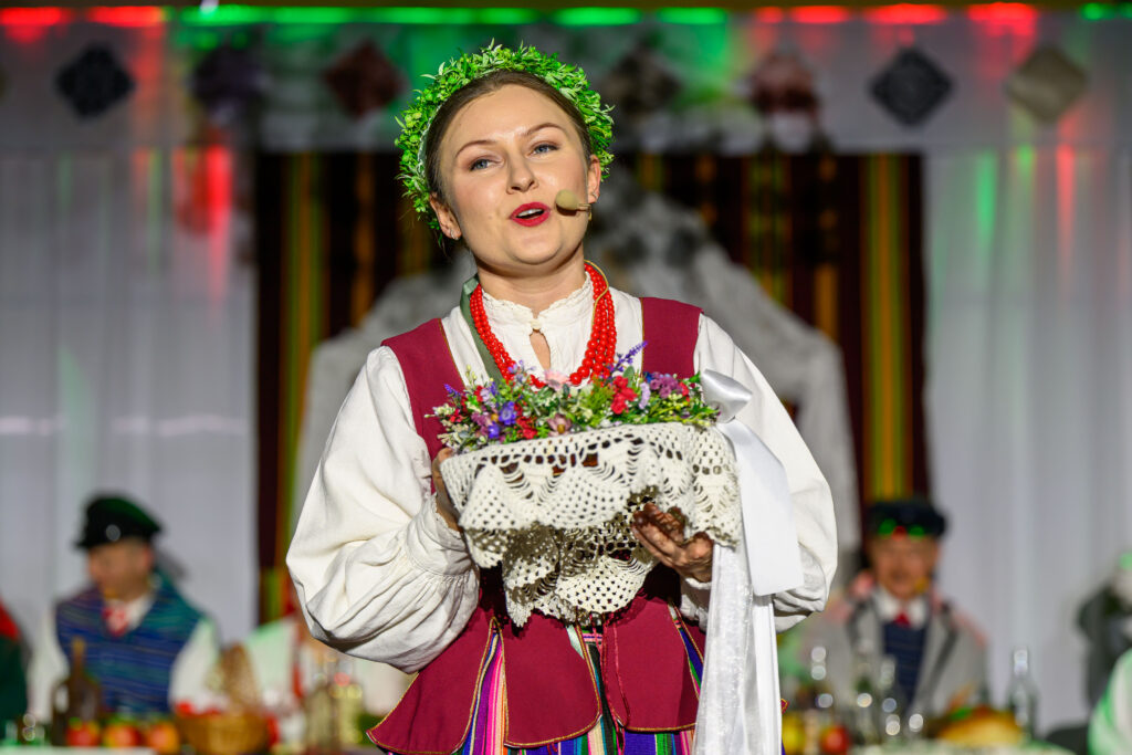 Kobieta w stroju ludowym z wieńcem na głowie trzyma koszyk z kwiatami. Tło barwne i rozmyte. Wyraźne kolory: biały, czerwony.