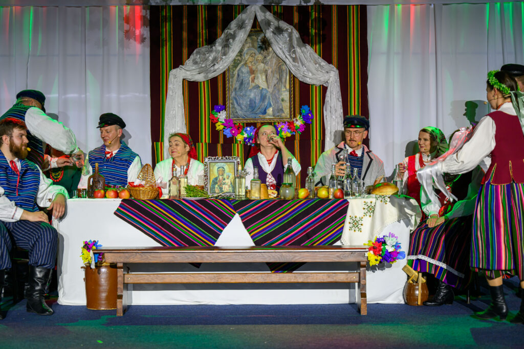Grupa osób w tradycyjnych strojach ludowych siedzi przy stole nakrytym kolorowym obrusem. Tło zdobią białe zasłony i obraz religijny w otoczeniu kwiatów. Na stole znajdują się jabłka, chleb i napoje.