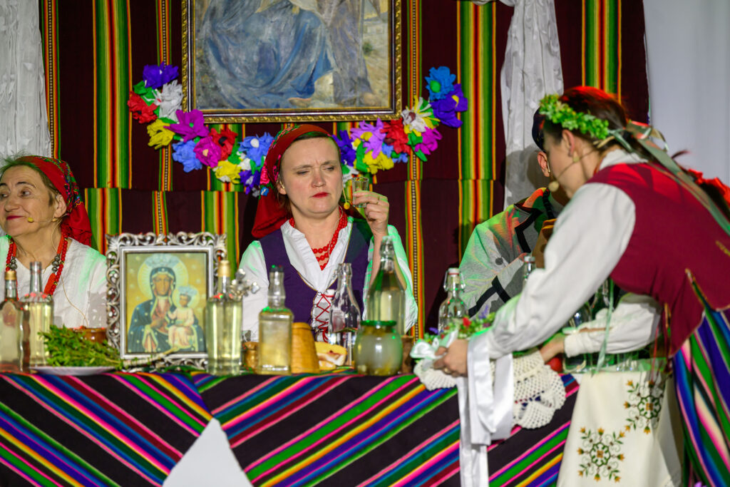 Na zdjęciu widać osoby w strojach ludowych przy stole. Tło zdobią kolorowe pasy i kwiatowe dekoracje. Na stole stoją butelki i obraz Matki Boskiej.