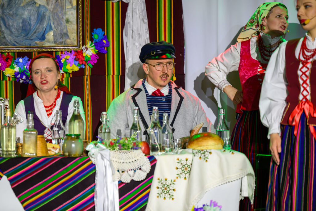 Na zdjęciu są osoby w strojach ludowych przy stole z kolorowym obrusem. Tło zdobią kwiaty i obrazy. Atmosfera jest barwna i kulturalna.