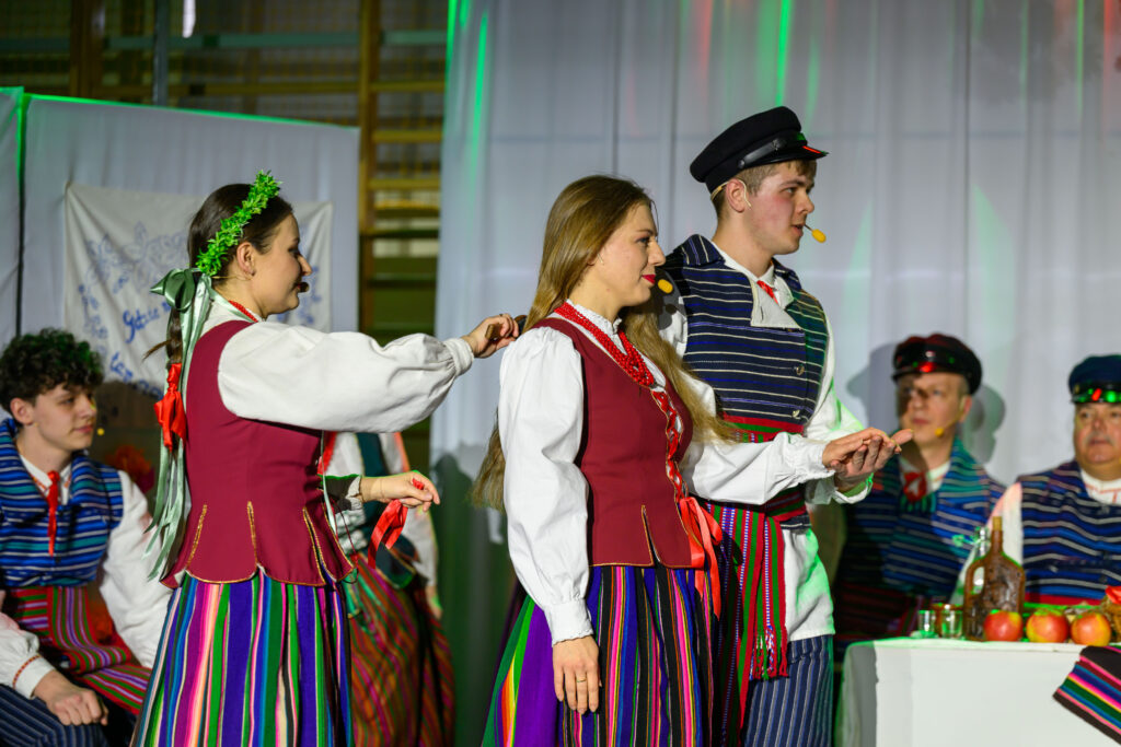 Trzy osoby w kolorowych strojach ludowych tańczą na scenie. Tło stanowi biała zasłona z delikatnym oświetleniem. Na stole obok widoczne są jabłka i karafka.