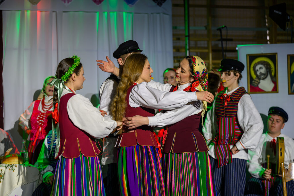 Grupa osób w tradycyjnych, kolorowych strojach ludowych tańczy na scenie. Tło jest jasne, ozdobione obrazami religijnymi. Scena jest żywa, przeważają barwy czerwone, zielone i białe.