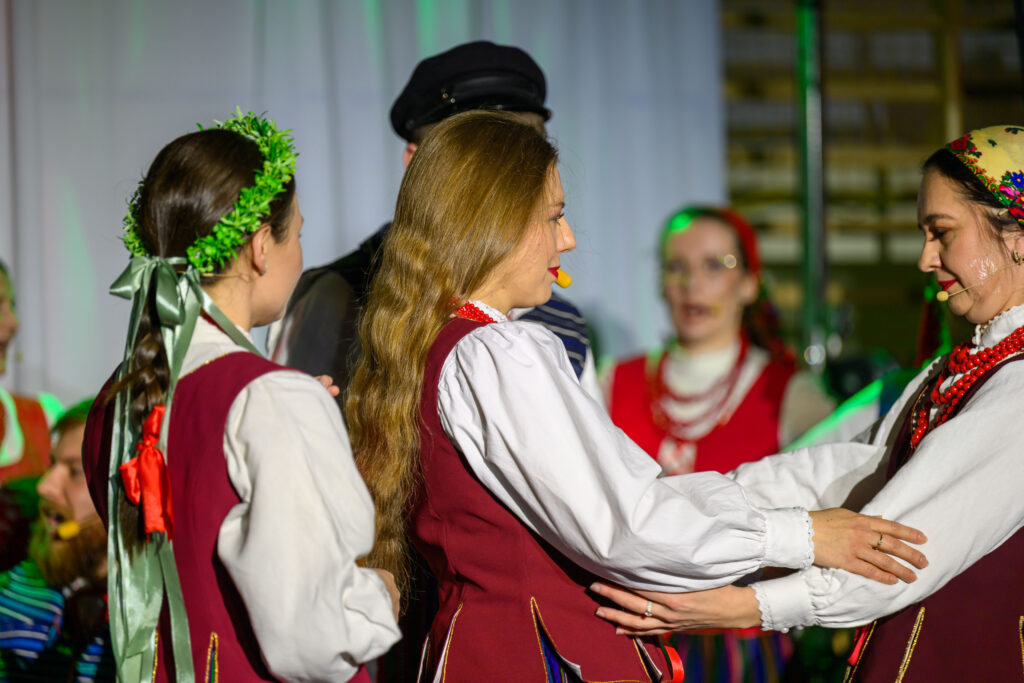 Grupa kobiet w strojach ludowych tańczy na scenie. Mają czerwone kamizelki i białe koszule, tło jest jasne. Panuje radosna atmosfera.